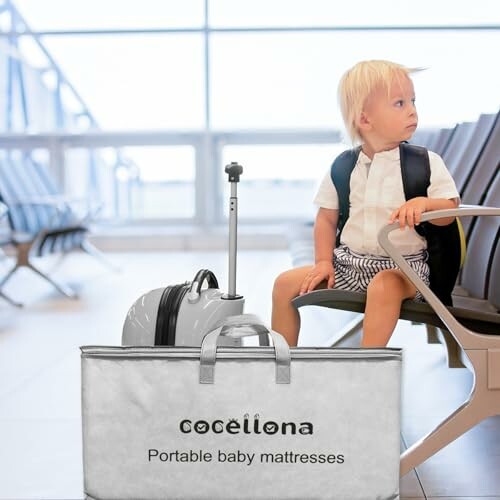 Toddler sitting on airport chair with luggage and portable baby mattress.